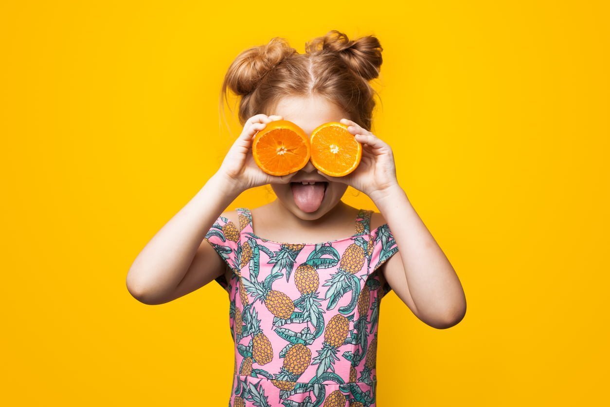 Caucasian Small Girl With Blonde Hair And Summer Dress Is Posing With Sliced Oranges On Eyes And Opened Mouth On A Yellow Studio Wall